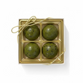 A square white and gold gift box containing four round green speckled pastries or candies, photographed among other similar packaged treats.